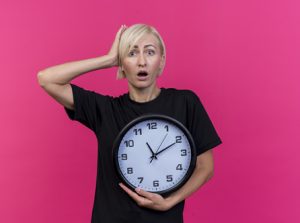 surprised-middle-aged-blonde-slavic-woman-looking-camera-holding-clock-putting-hand-head-isolated-crimson-background-with-copy-space2